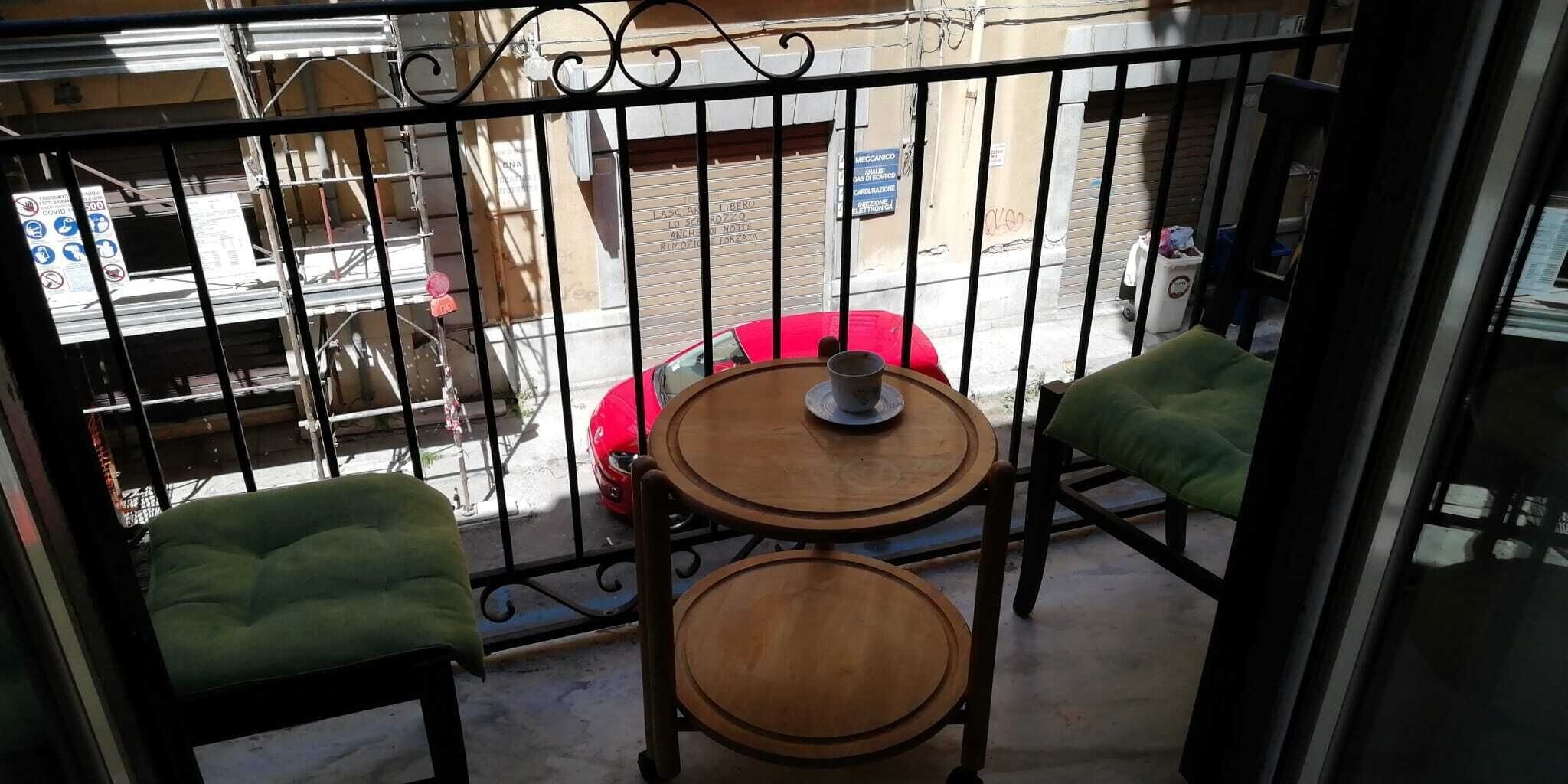 A balcony with two wooden tables and green cushioned chairs, overlooking a street with a red car and a closed shuttered building.