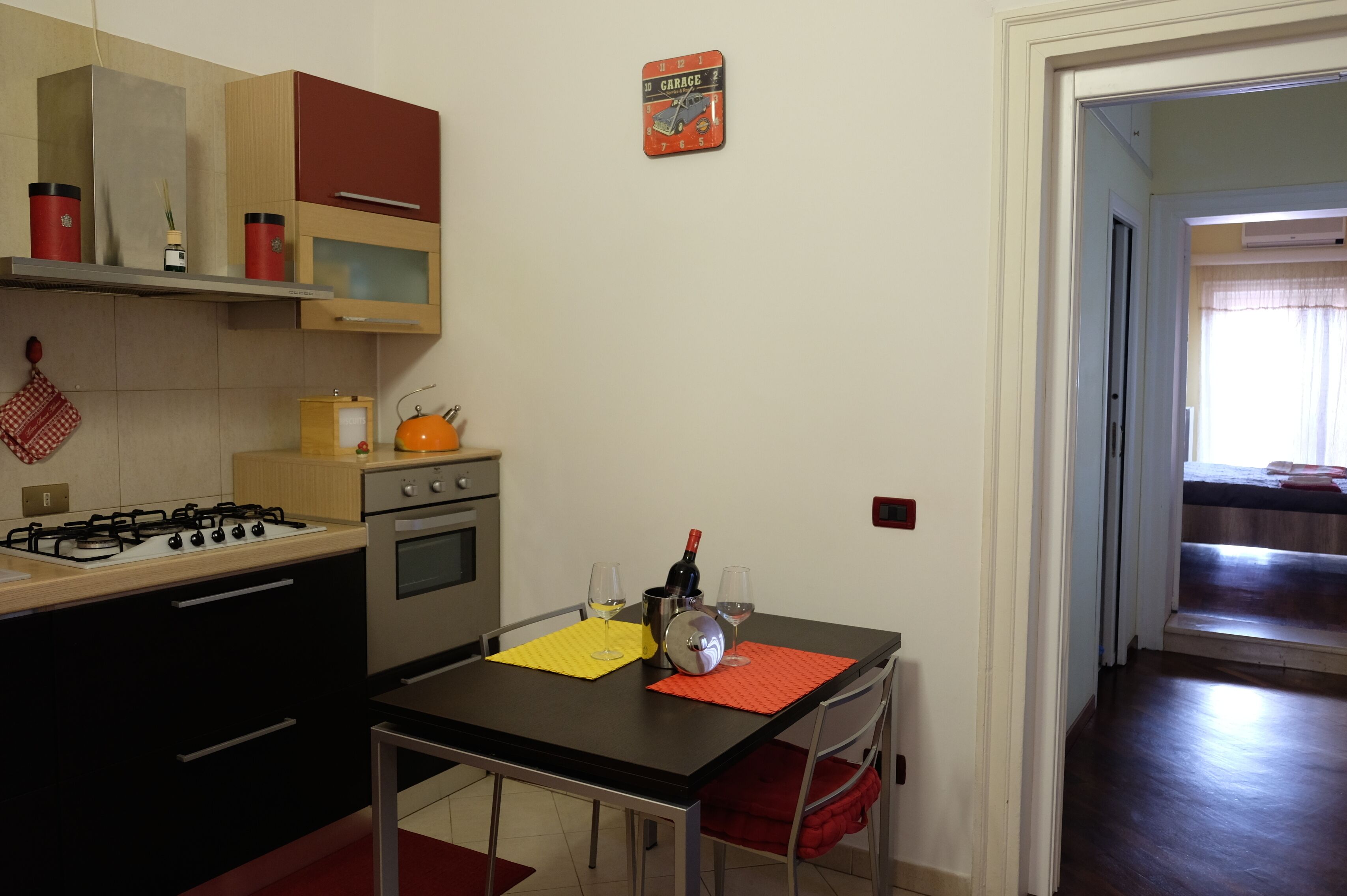 Cocina con estufa a gas, horno y microondas. Hay una mesa con dos copas de vino y una botella. Un mantel individual rojo y amarillo está sobre la mesa.