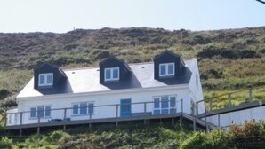 A two-story house with a white exterior and dark roof, situated on a hillside with a view of the surrounding landscape. - Contemporary Beach House with sea views and short walk to Blue Flag beach (Porthtowan)