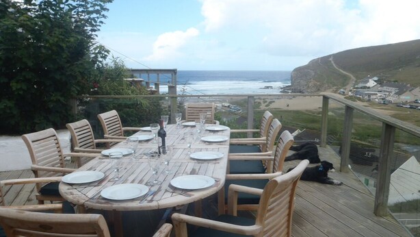 A wooden deck with a round table set for dining, overlooking a beach and ocean. - Contemporary Beach House with sea views and short walk to Blue Flag beach (Porthtowan)