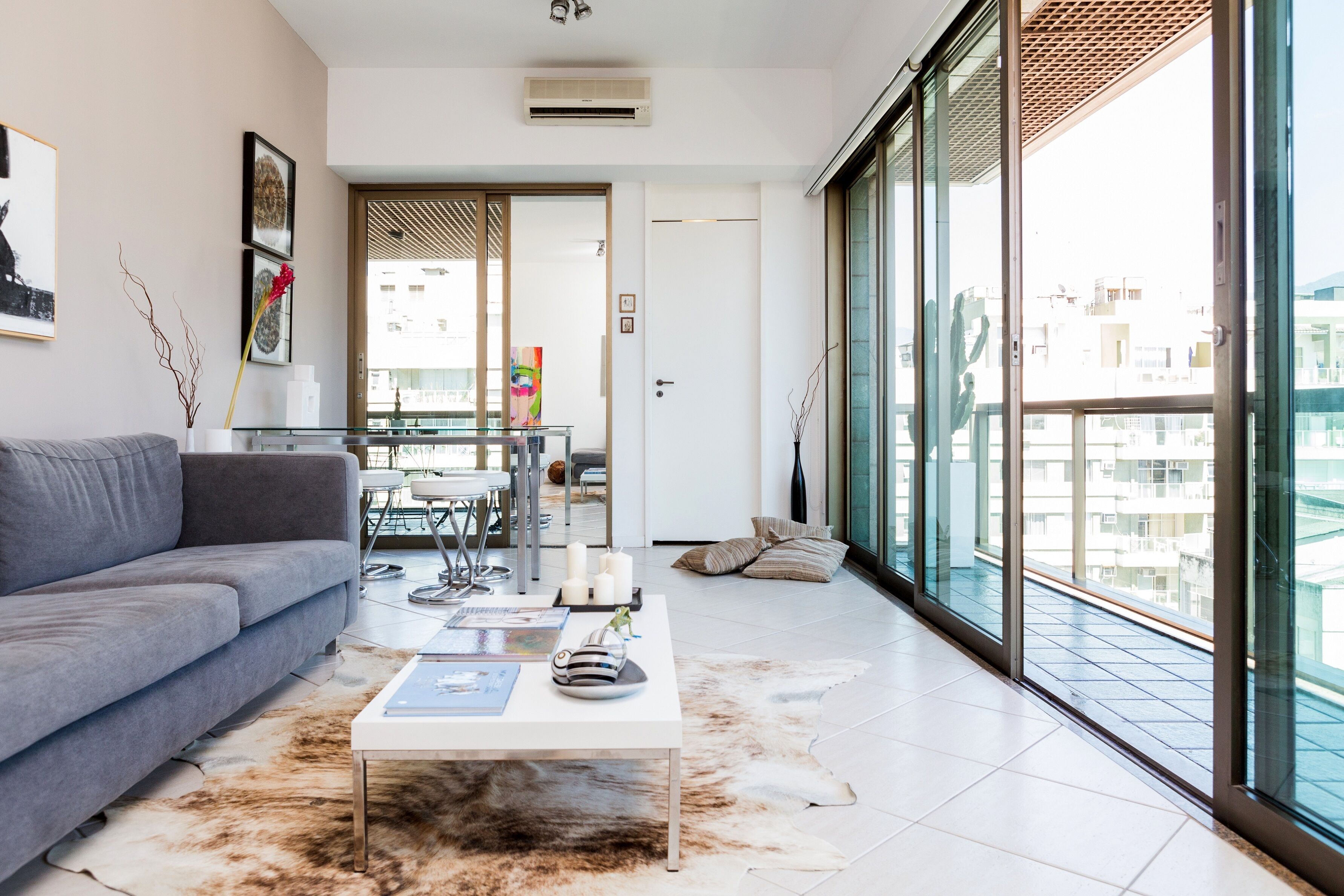 Uma sala de estar moderna com um sofá cinzento, uma mesa de centro de vidro e uma mesa de jantar de vidro com cadeiras. Há uma janela grande com vista para edifícios.