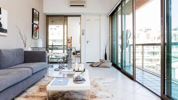 Uma sala de estar moderna com um sofá cinzento, uma mesa de centro de vidro e uma mesa de jantar de vidro com cadeiras. Há uma janela grande com vista para edifícios.
