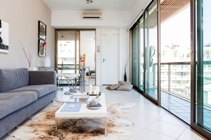 A modern living room with a grey sofa, a glass coffee table, and a glass dining table with chairs. There is a large window with a view of buildings outside. - Heart of Ipanema, stylish apartment in full service building. IDEAL LOCATION! (Rio de Janeiro)