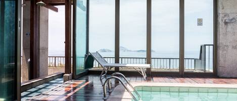 A pool area with a view of the sea, a lounge chair, and a small table.