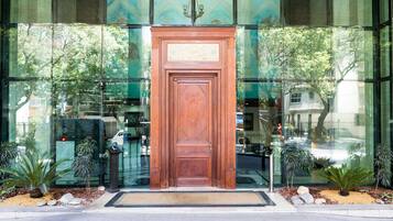 Uma porta de madeira com um painel decorativo, ladeada por janelas de vidro que refletem árvores e um edifício.