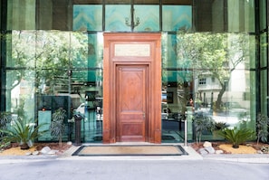 A wooden door with a decorative panel, flanked by glass windows reflecting trees and a building. - Heart of Ipanema, stylish apartment in full service building. IDEAL LOCATION! (Rio de Janeiro)