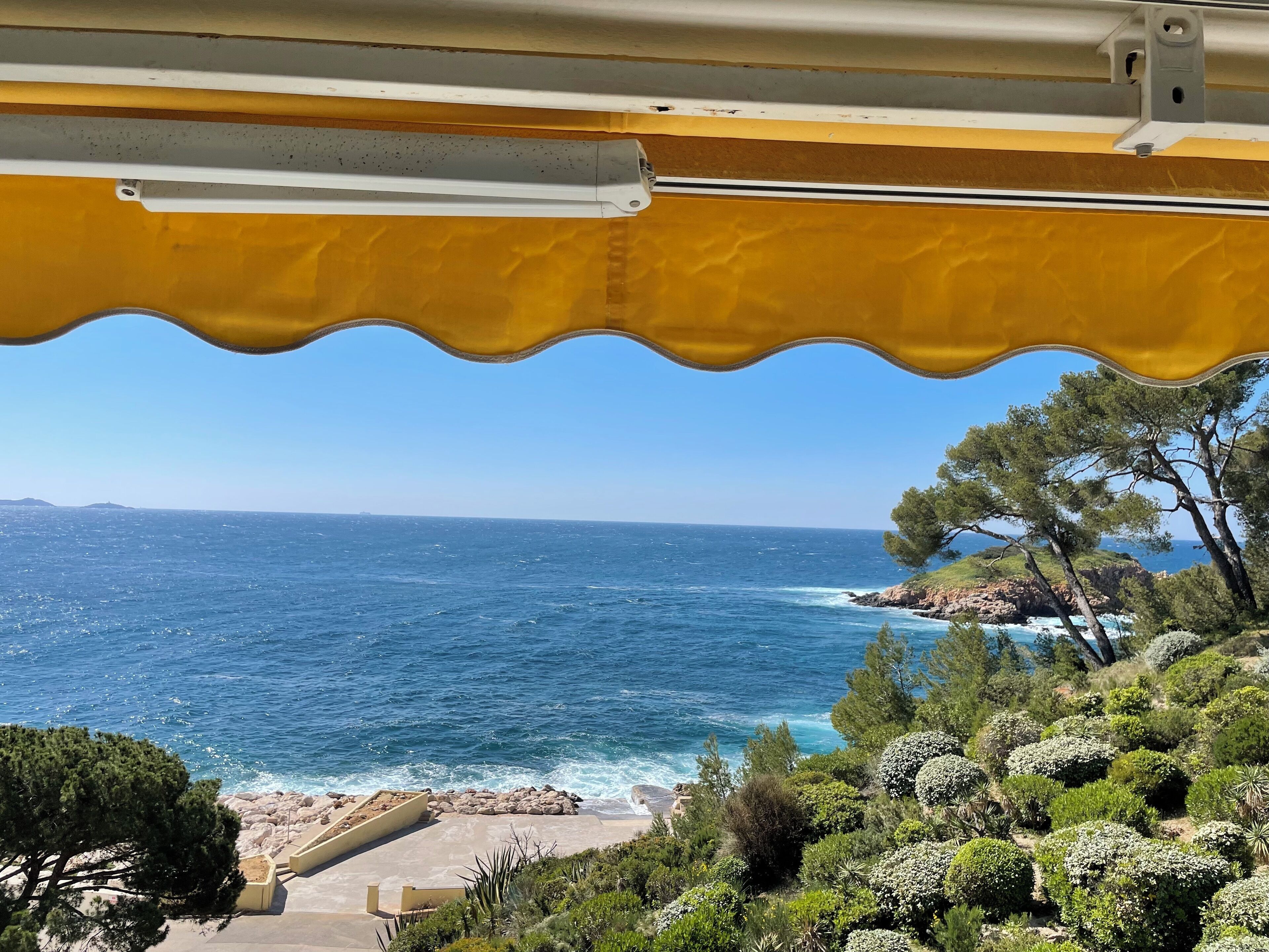 Une vue sur la côte, depuis une fenêtre surmontée d’un auvent jaune, donnant sur la mer et un paysage rocheux parsemé d’arbres.