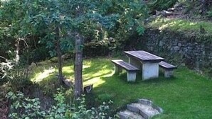 Eine Stehbank und ein Picknicktisch in einer grünen Parklandschaft mit Bäumen und einer Steinmauer.