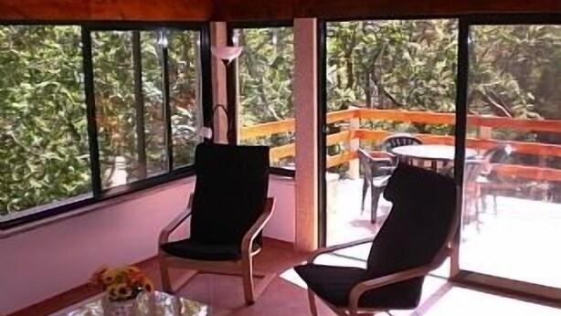 Ein Zimmer mit zwei Sesseln, die zu einem großen Fenster zeigen, einem kleinen Tisch mit Glasplatte und Blick auf eine Außenterrasse mit Holzgeländer.