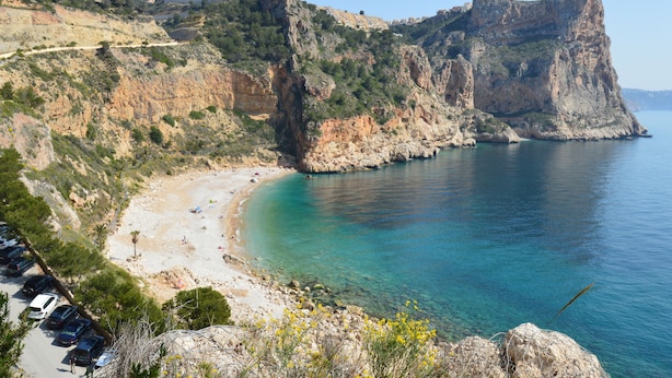 Vista costera con un acantilado rocoso, mar azul cristalino y una playa de arena.