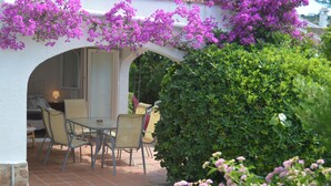 Un patio al aire libre con una mesa y sillas, rodeado de abundante follaje y plantas con flores.
