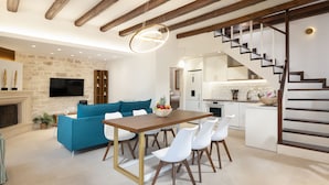 A modern living room with a blue sofa, a wooden dining table, and a staircase leading to a kitchen. - Dafni Villa, a Blissful Retreat with 40m² Pool (Rethymno)