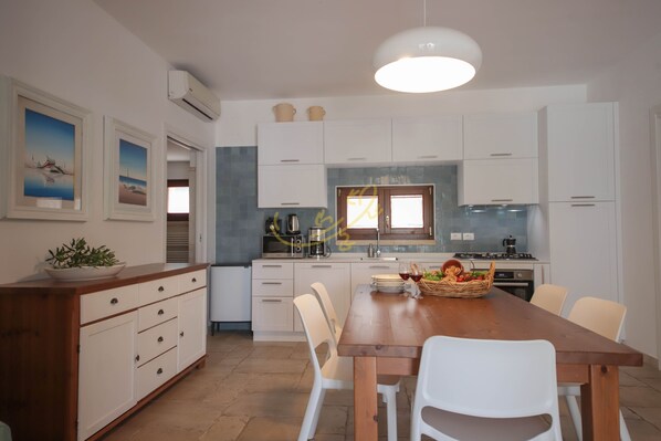 Une cuisine moderne avec des armoires blanches, une table à manger en bois et des chaises blanches. Il y a une fenêtre donnant sur l’extérieur et un luminaire au plafond.
