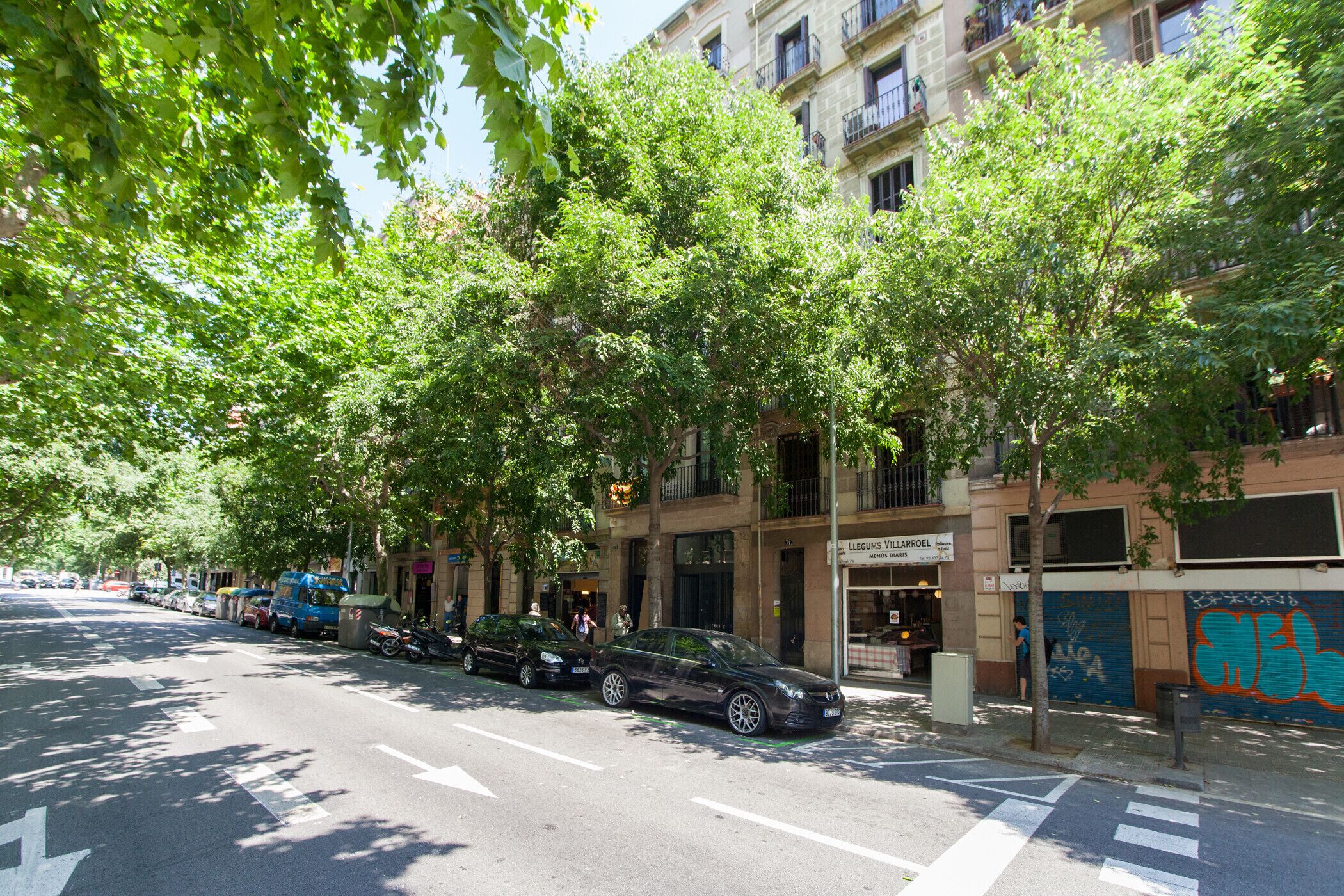 Una calle bordeada de árboles y automóviles estacionados, con un edificio que tiene un restaurante llamado 'Legumes Villarroel' en la planta baja.