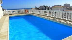 Una piscina rettangolare con ringhiera bianca, un galleggiante salvagente e vista sul mare e sugli edifici sullo sfondo.