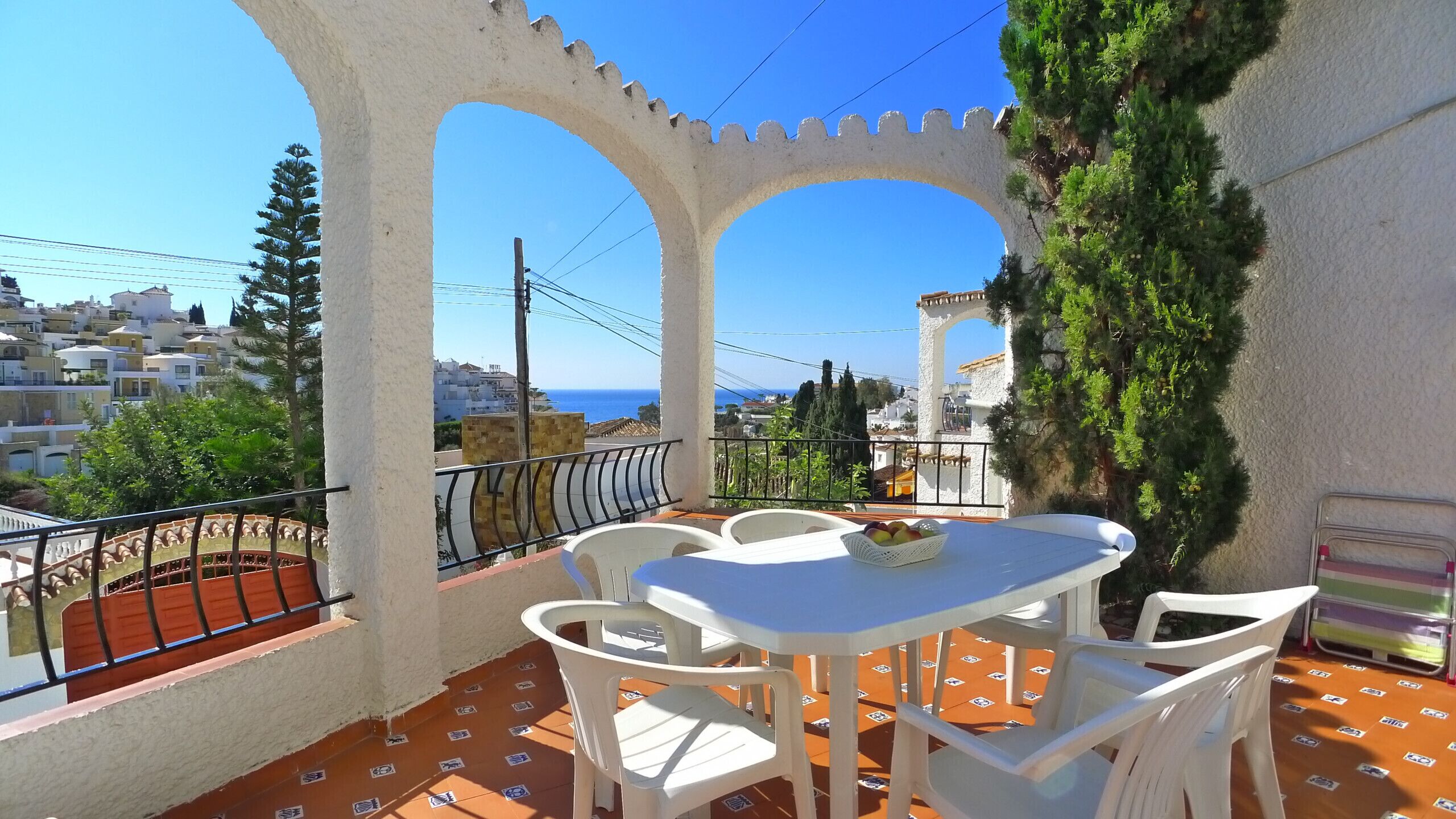 A terrace with a table and chairs, a view of the sea, and a white building with arches.