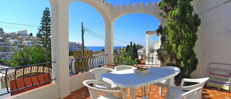 A terrace with a table and chairs, a view of the sea, and a white building with arches.