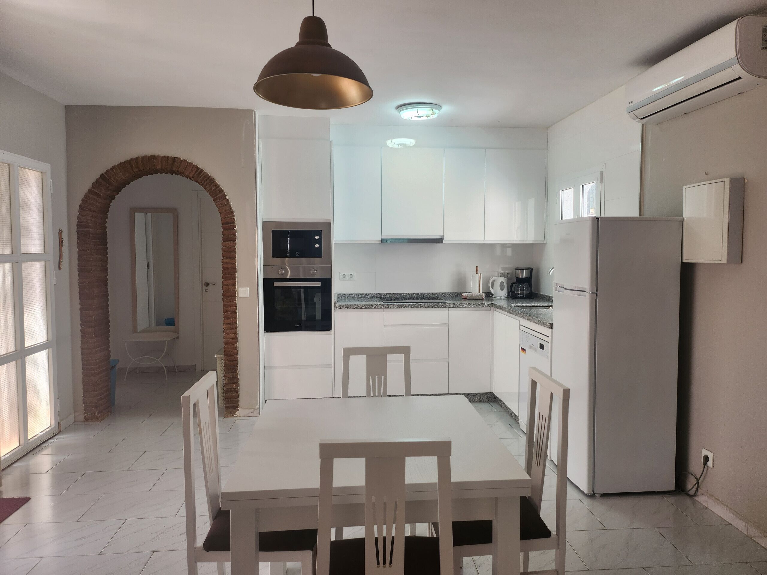Une cuisine moderne avec des meubles blancs, une table à manger avec des chaises, un four, un réfrigérateur et un luminaire suspendu.