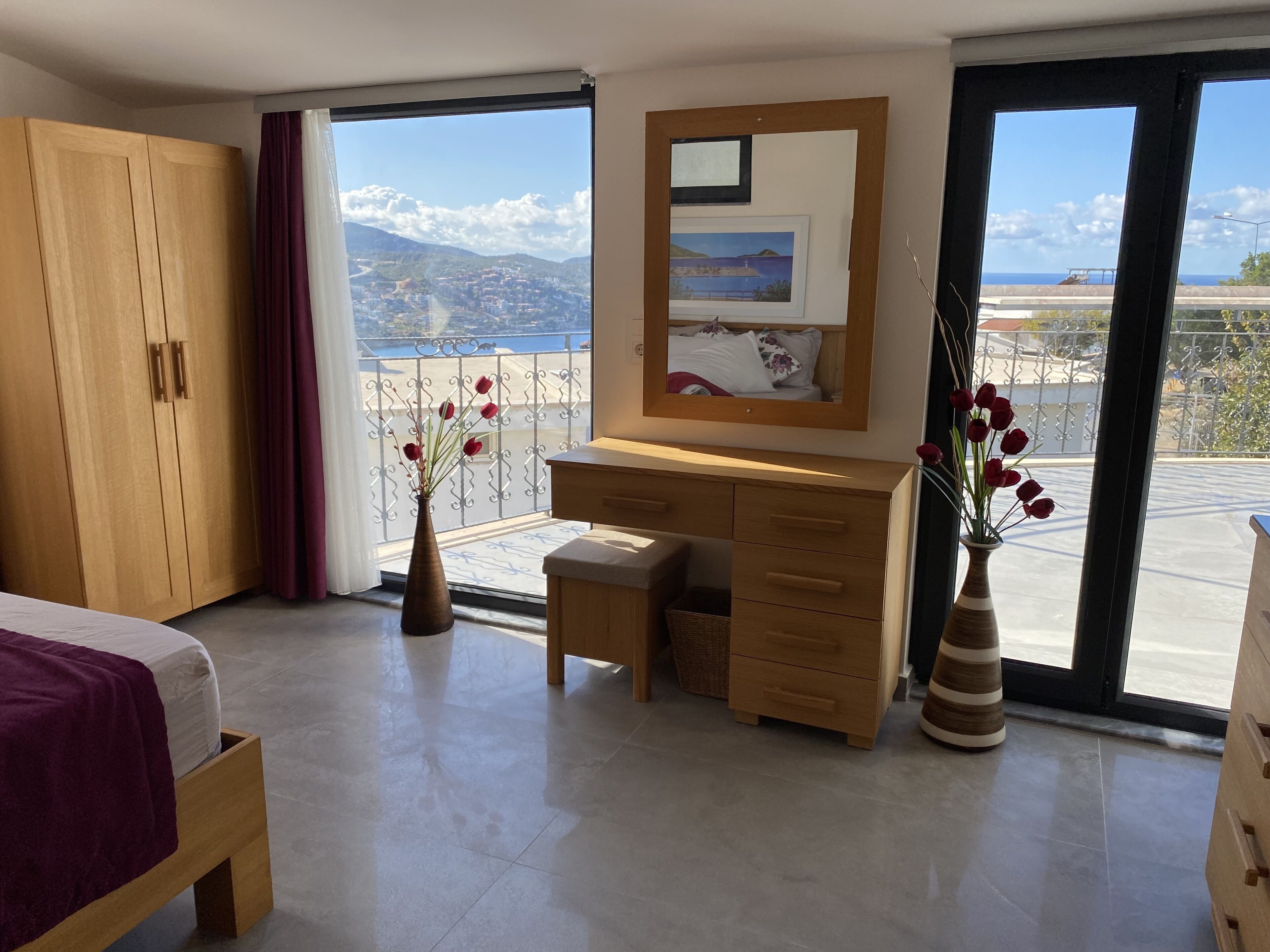 A modern bedroom with a large window, wooden wardrobe, desk, and bedside table.