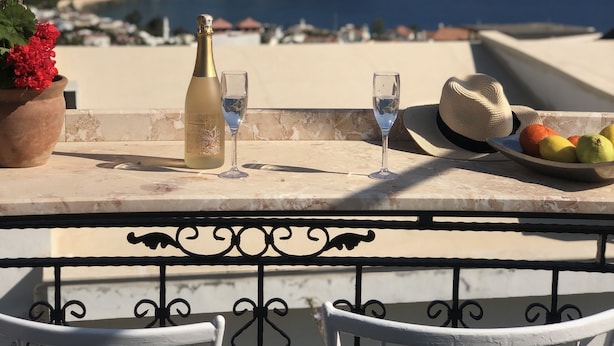 Een terras met een tafel gedekt voor twee personen, met een fles, glazen, een hoed en een fruitschaal. Het terras heeft uitzicht op een kustlandschap met gebouwen en heuvels.