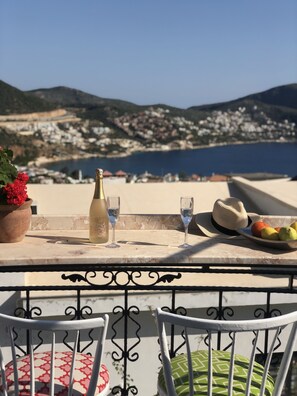 En terrasse med et bord dækket til to personer, der inkluderer en flaske, glas, en hat og en skål med frugt. Terrassen har udsigt over et kystlandskab med bygninger og bakker.