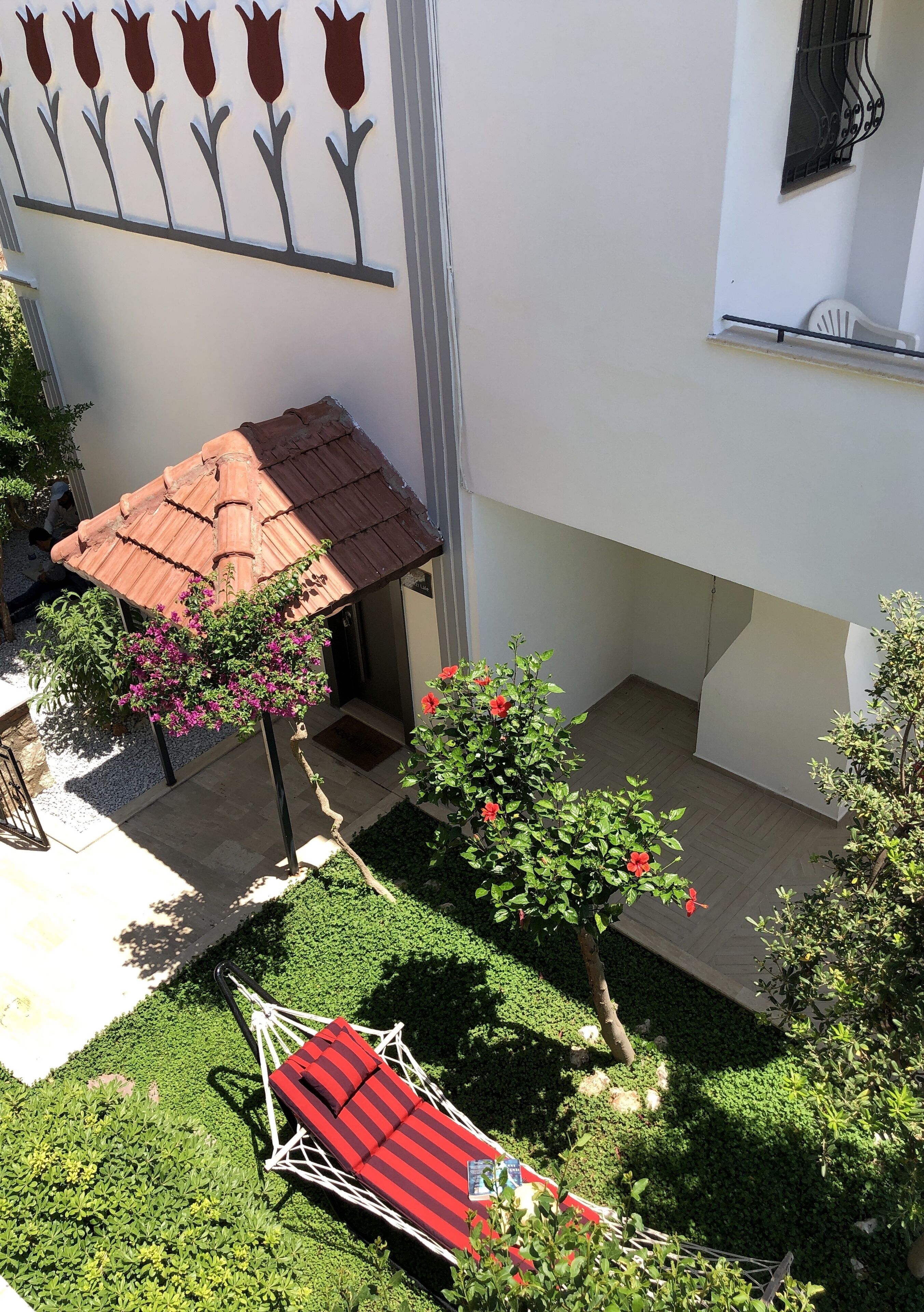 Ein kleiner Außenbereich mit einer rot-gestreiften Hängematte, einer Dachkonstruktion mit Ziegeldeckung und einer an der Wand befestigten Blumendekoration.
