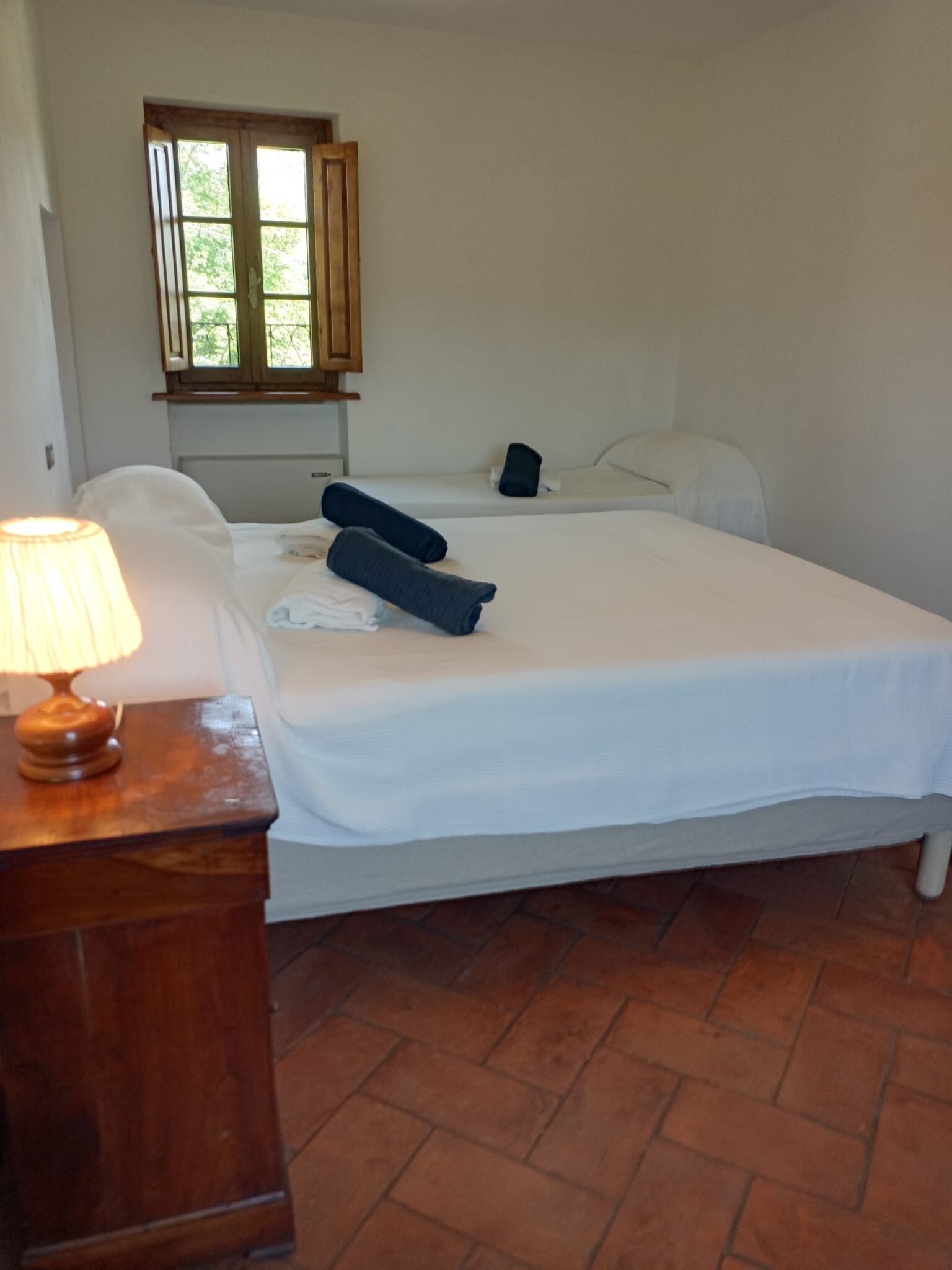Une chambre avec un lit simple, une table de chevet en bois, une lampe, une fenêtre avec des volets en bois et un sol carrelé.