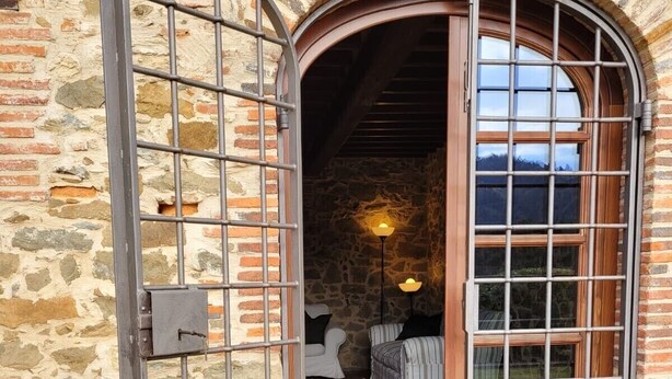 Un portail en arc surmonté d’une grille métallique, qui donne sur une pièce avec un mur en pierre, un lit et une lampe.