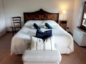 Een net opgemaakt bed met een gestreept kussen en opgevouwen kleding erop, een houten stoel, een nachtkastje met een lamp, en een klein wit apparaat.