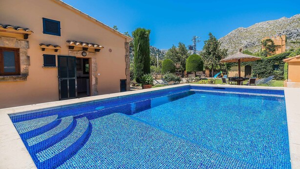 Une piscine aux carreaux de mosaïque bleus, un bâtiment à la façade beige et un paysage montagneux en arrière-plan.