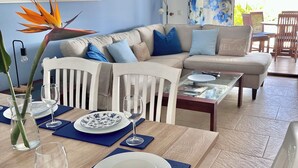 A living room with a sofa, dining table set with plates and glasses, a white chair, and a large window with curtains.