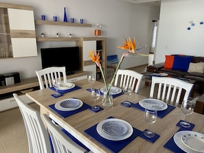 Un comedor con una mesa de madera dispuesta para cuatro, sillas blancas y manteles individuales azules.
