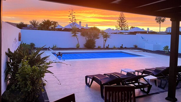A pool with lounge chairs, a covered area, and a view of a sunset over a white-walled building.