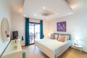 A bedroom with a bed, a TV, a desk with a bottle, a mirror, and a window with curtains.