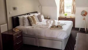 Una cama bien hecha con un cojín con forma de cisne, una mesita de noche con lámpara y una ventana con cortinas.
