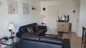 Una sala de estar con un sofá negro, una mesa de centro de vidrio, una lámpara y un televisor sobre un mueble de madera.