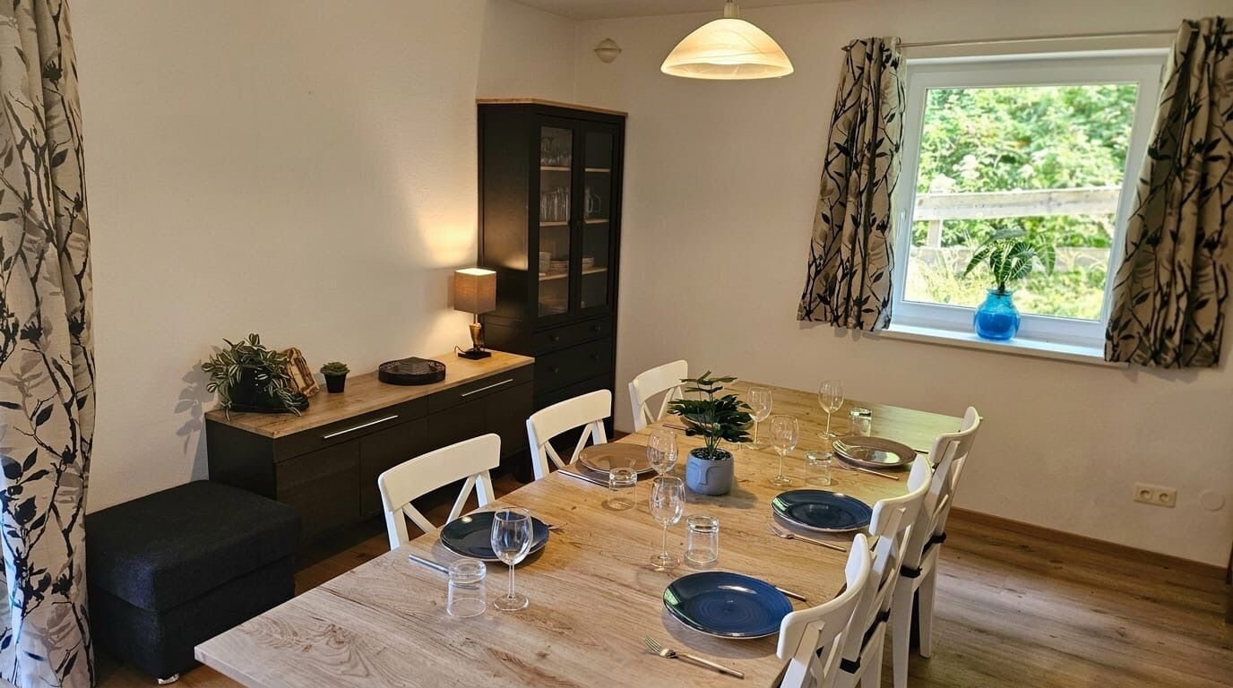 Un coin repas avec une table en bois dressée pour quatre personnes, des chaises blanches, un buffet, une lampe et une fenêtre donnant sur un paysage verdoyant.