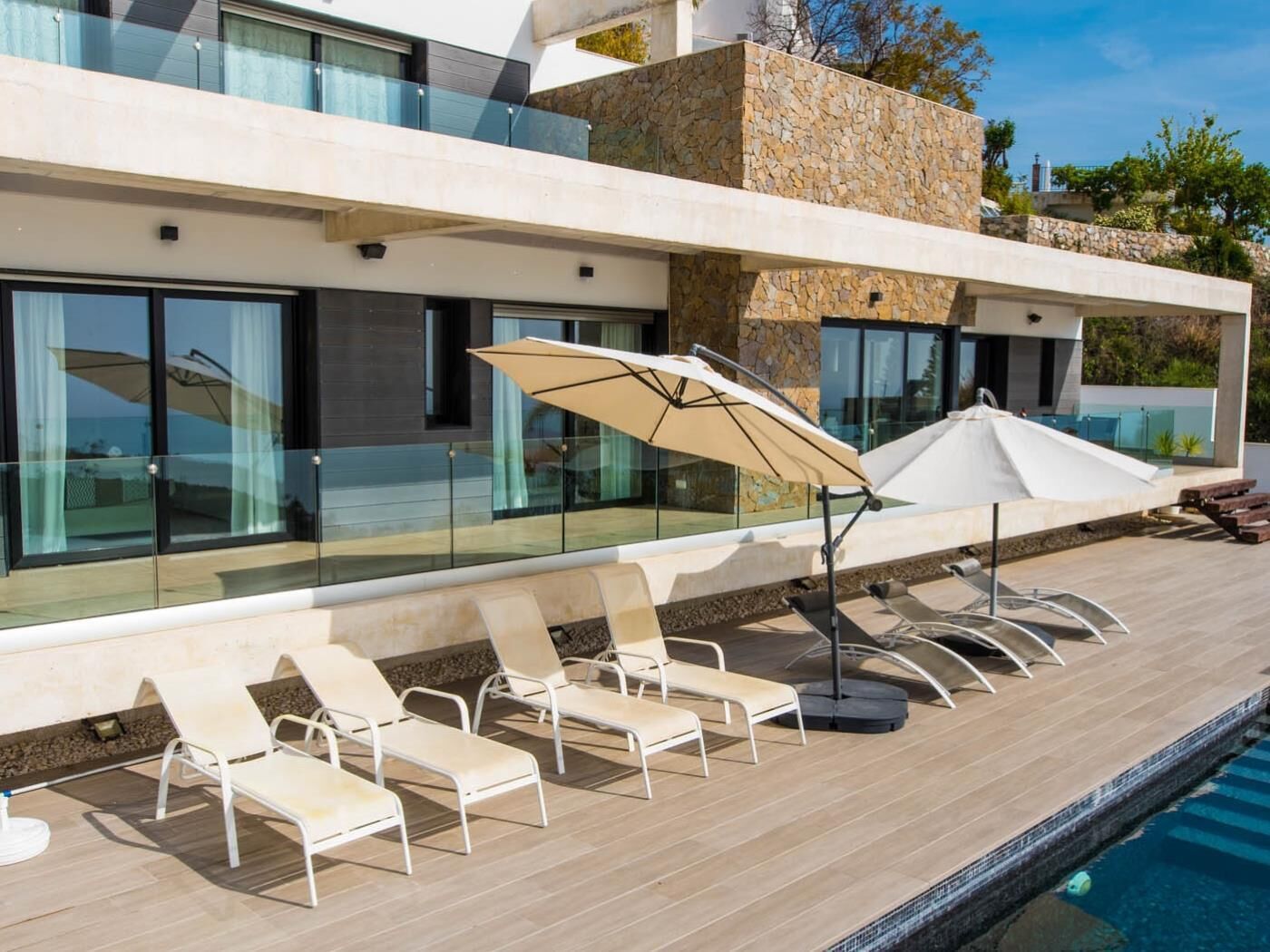 A modern poolside area with lounge chairs and umbrellas, large glass windows, and a stone facade.