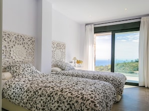 Two beds with patterned bedspreads, a large window offering a view of the landscape and sea, and a decorative wall panel.