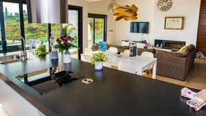 A modern kitchen with a central island, dining area with white chairs, a brown sofa, and a TV mounted on the wall. - Luxury villa with heated pool and sea views (Salobreña)