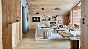 A cozy, wooden-floored living area with a sofa, coffee table, and chairs, a bookshelf, and a large window. - Barma Chalet - Luxurious modern chalet, exceptional view, 900 m from the slopes (Manigod)