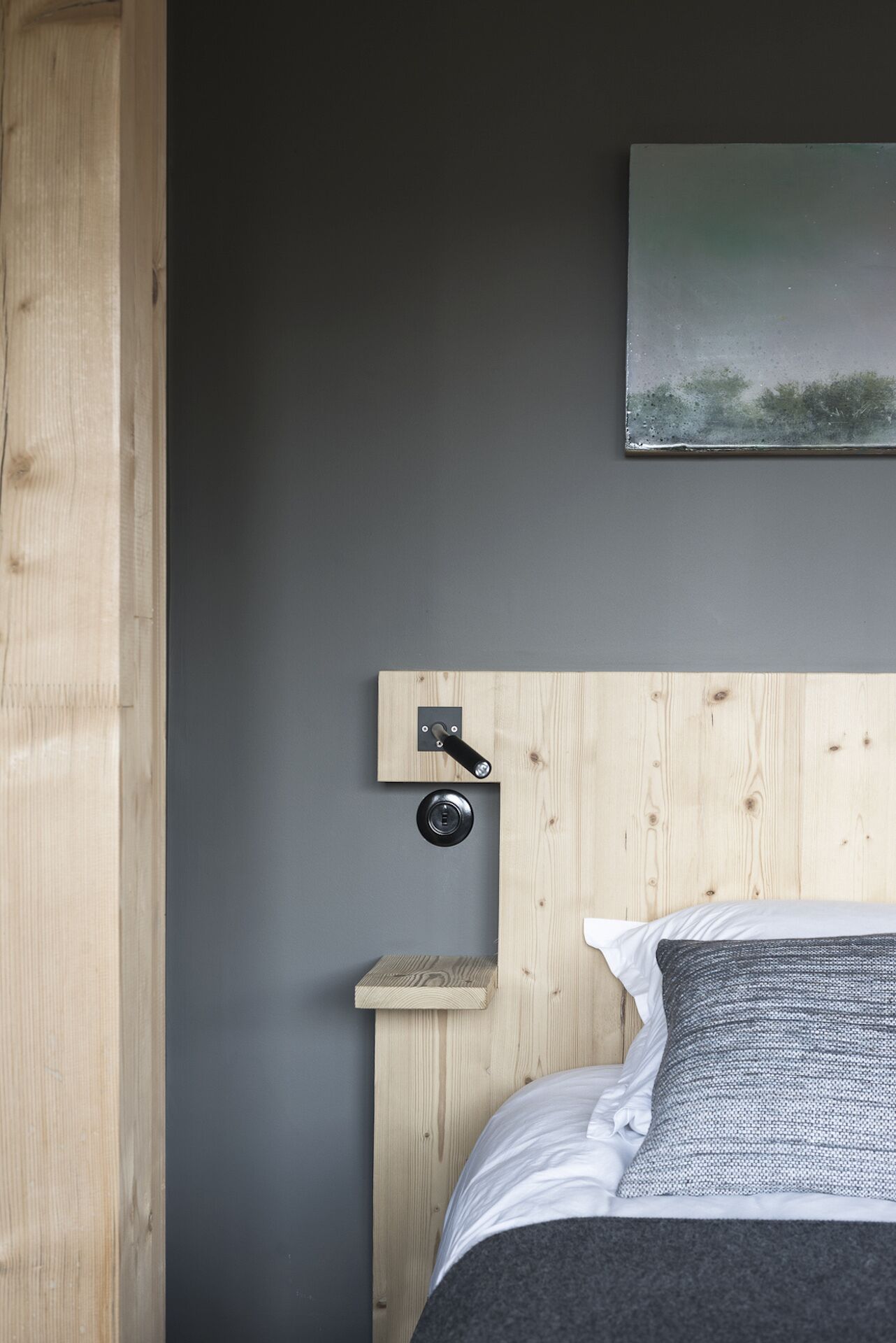 Un dormitorio con cabecera de madera, un estante pequeño de madera, una mesita de noche redonda y negra, una cama con ropa de cama blanca y gris, y una pared gris.