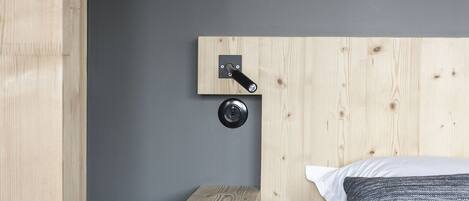Un dormitorio con cabecera de madera, un estante pequeño de madera, una mesita de noche redonda y negra, una cama con ropa de cama blanca y gris, y una pared gris.