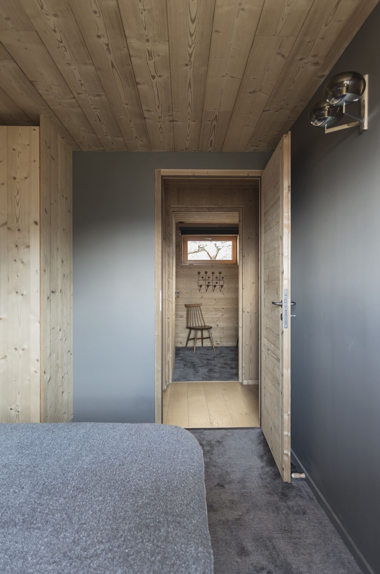 A room with a wooden ceiling, a grey carpet, a small wooden table with a chair, and a window with a view of trees.