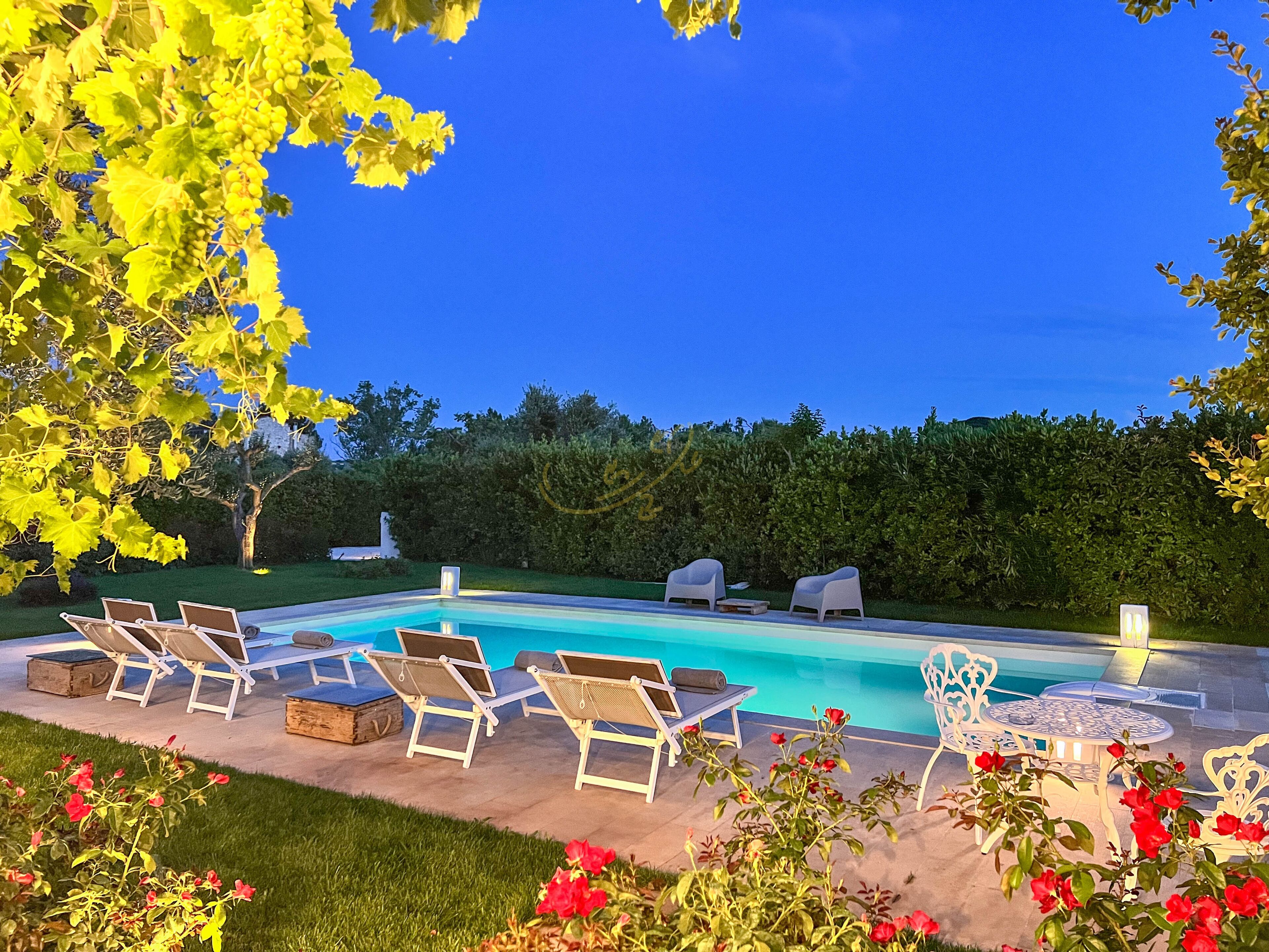 Ein Pool mit Liegestühlen und einem Tisch, umgeben von Grünpflanzen und Blumen.