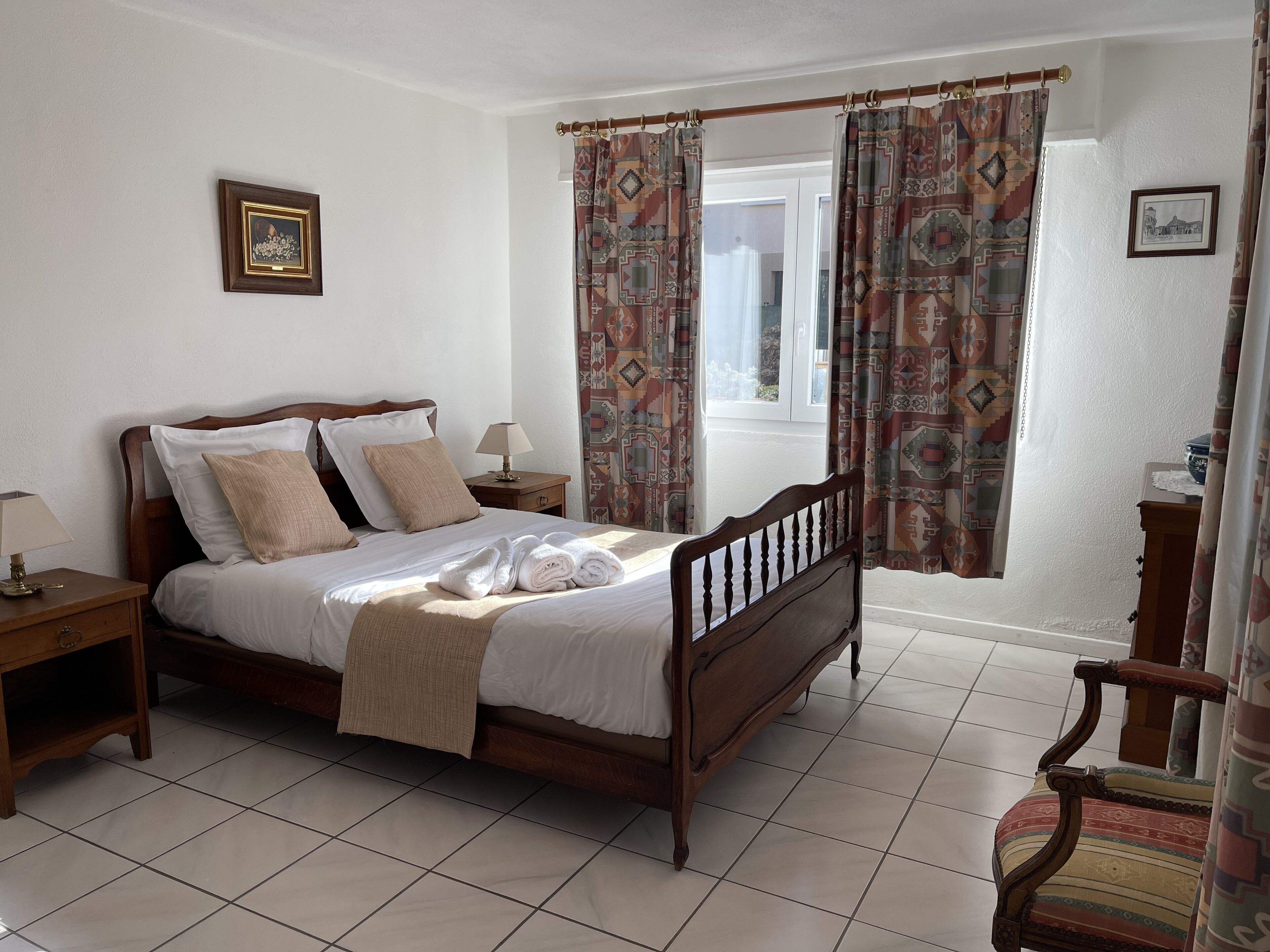 A neatly made bed with a wooden headboard, a nightstand with a lamp, and a window with patterned curtains.