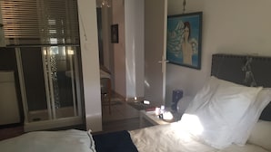Quarto de hotel com cama, mesa de cabeceira com abajur, quadro na parede e porta deslizante para uma varanda.