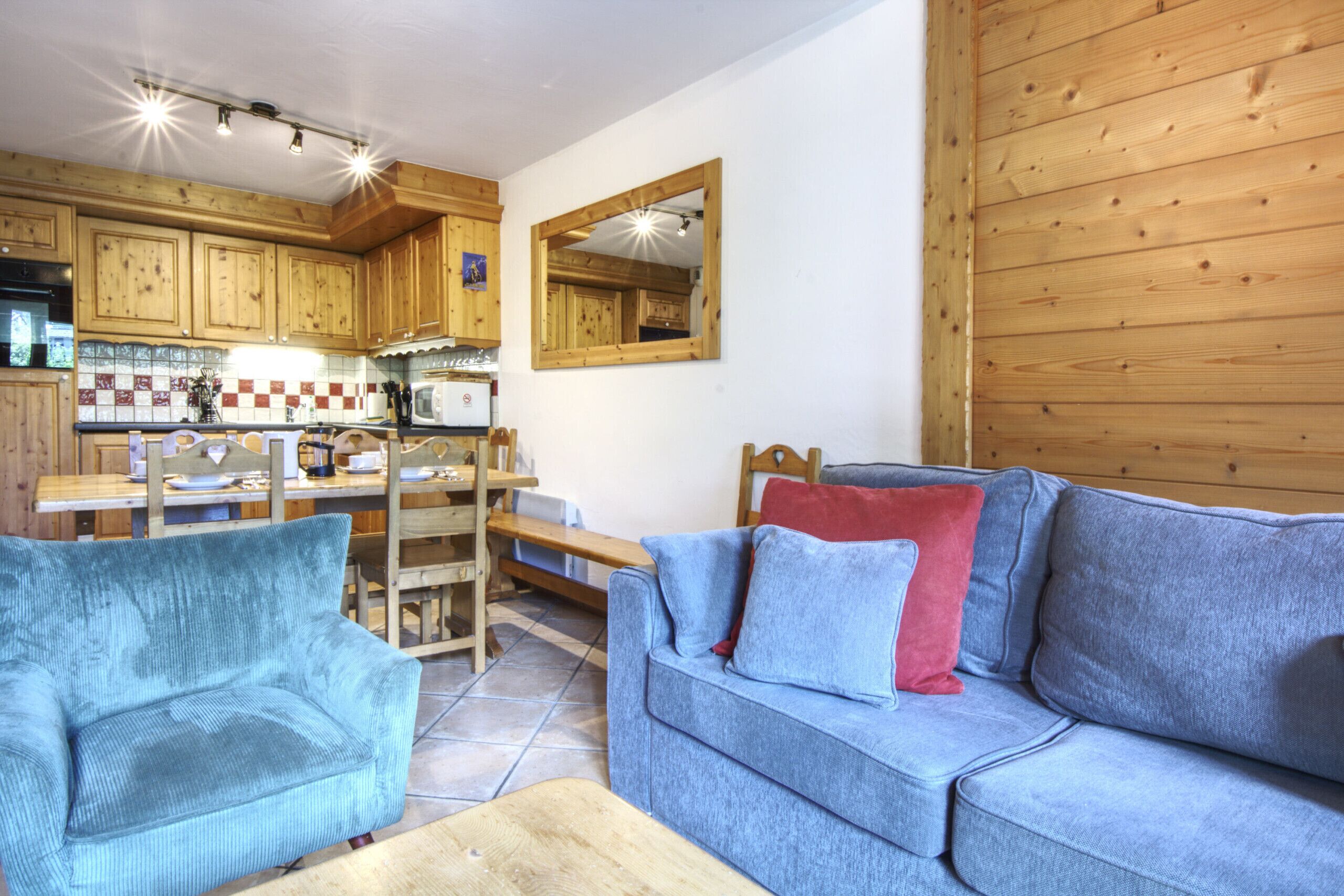 Un salon chaleureux avec un canapé bleu, un fauteuil assorti et une table à manger en bois avec des chaises. On aperçoit, au fond, une cuisine avec des meubles en bois et un four à micro-ondes.