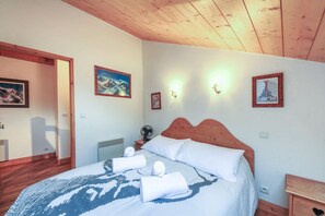 Camera da letto con soffitto in legno, un letto con lenzuola bianche e una coperta fantasia, un comodino con una lampada e luci a parete.