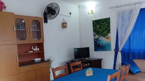 A dining area with a blue tablecloth, wooden chairs, a TV, and a painting of a beach scene on the wall.